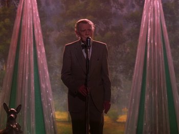 Movie still from “Twin Peaks” (1990), created by David Lynch – A man standing in front of a microphone in front of trees; Medium shot, Low angle