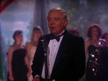 Movie still from “Twin Peaks” (1990), created by David Lynch – An older man in a tuxedo is speaking into a microphone in front of a crowd; Medium shot, Over the shoulder angle