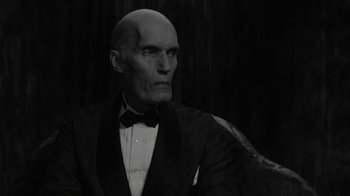 Movie still from “Twin Peaks” (2017), directed by David Lynch – A bald man in a suit and bow tie looking to his left; Medium shot, Over the shoulder angle