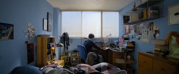 Movie still from “Anonymously Yours” (2021), directed by Maria Torres – A man sitting at a desk in front of a large window; Wide shot, High angle