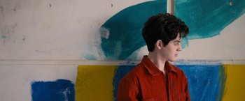 Movie still from “Anonymously Yours” (2021), directed by Maria Torres – A man in a red shirt is standing in front of a painting; Close Up shot, Over the shoulder angle