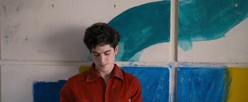 Movie still from “Anonymously Yours” (2021), directed by Maria Torres – A young man in a red shirt is standing in front of a painting; Medium shot, Over the shoulder angle