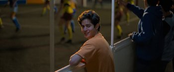 Movie still from “Anonymously Yours” (2021), directed by Maria Torres – A young man leaning on a railing in front of a crowd of soccer players; Medium shot, Over the shoulder angle