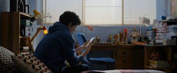 Movie still from “Anonymously Yours” (2021), directed by Maria Torres – A young man is sitting on a bed and using a tablet; Medium shot, High angle