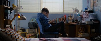 Movie still from “Anonymously Yours” (2021), directed by Maria Torres – A young man sitting in a chair looking at his phone; Medium shot, High angle