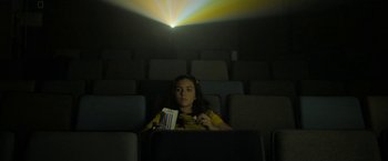 Movie still from “Anonymously Yours” (2021), directed by Maria Torres – A woman sitting in a chair in a movie theater holding a book; Medium shot, Low angle