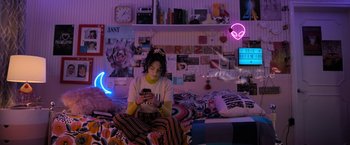 Movie still from “Anonymously Yours” (2021), directed by Maria Torres – A woman sitting on top of a bed looking at her phone; Wide shot, High angle