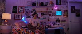 Movie still from “Anonymously Yours” (2021), directed by Maria Torres – A woman sitting on top of a bed in a room; Wide shot, High angle