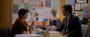 Movie still from “Anonymously Yours” (2021), directed by Maria Torres – Two people sitting at a table in front of papers and pens; Medium shot, Over the shoulder angle