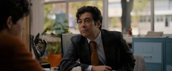 Movie still from “Anonymously Yours” (2021), directed by Maria Torres – A man sitting at a table wearing a suit and tie; Close Up shot, Over the shoulder angle