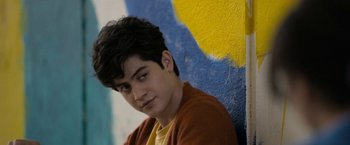 Movie still from “Anonymously Yours” (2021), directed by Maria Torres – A young man is posing in front of a colorful wall; Close Up shot, Over the shoulder angle