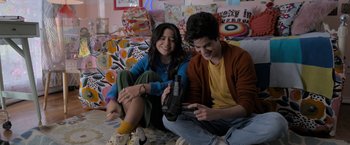 Movie still from “Anonymously Yours” (2021), directed by Maria Torres – Two young people sitting on the floor looking at an electronic device; Medium shot, Over the shoulder angle