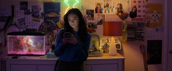 Movie still from “Anonymously Yours” (2021), directed by Maria Torres – A woman standing in a kitchen looking at her phone; Medium shot, High angle