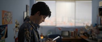 Movie still from “Anonymously Yours” (2021), directed by Maria Torres – A man looking at his cell phone while standing in front of a window; Medium shot, Over the shoulder angle