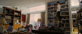 Movie still from “Anonymously Yours” (2021), directed by Maria Torres – A man standing on top of a ladder in front of a bookshelf; Wide shot, Low angle