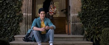 Movie still from “Anonymously Yours” (2021), directed by Maria Torres – A young man sitting on the steps of a house; Medium shot, Over the shoulder angle