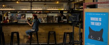Movie still from “Anonymously Yours” (2021), directed by Maria Torres – A woman sitting at a counter in front of a camera; Wide shot, High angle