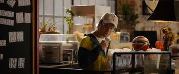 Movie still from “Anonymously Yours” (2021), directed by Maria Torres – A man wearing a baseball cap and glasses is eating a hot dog; Medium shot, Over the shoulder angle