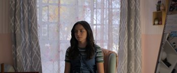 Movie still from “Anonymously Yours” (2021), directed by Maria Torres – A young girl sitting in a chair in front of a window; Medium shot, Over the shoulder angle