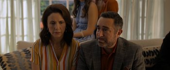 Movie still from “Anonymously Yours” (2021), directed by Maria Torres – A man and a woman sitting next to each other in front of other people; Close Up shot, Over the shoulder angle