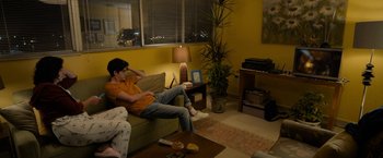 Movie still from “Anonymously Yours” (2021), directed by Maria Torres – A man sitting on top of a couch in a living room; Wide shot, High angle