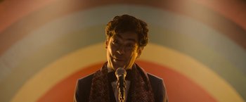 Movie still from “Anonymously Yours” (2021), directed by Maria Torres – A man with a microphone in front of a colorful background; Close Up shot, Low angle
