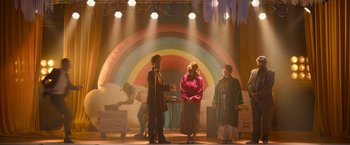 Movie still from “Anonymously Yours” (2021), directed by Maria Torres – A group of people standing on a stage with a rainbow in the background; Wide shot, Low angle