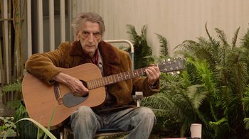 Movie still from “Twin Peaks” (2017), directed by David Lynch – An older man sitting in a chair playing a guitar; Medium shot, Over the shoulder angle