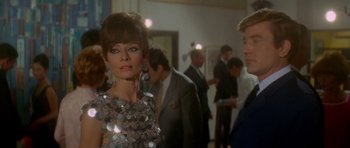 Movie still from “Two for the Road” (1967), directed by Stanley Donen – A woman standing next to a man in front of a crowd; Medium shot, Over the shoulder angle