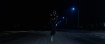 Movie still from “Two-Lane Blacktop” (1971), directed by Monte Hellman – A man standing in the middle of the road holding a skateboard; Wide shot, Low angle