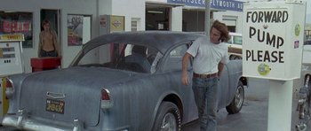 Movie still from “Two-Lane Blacktop” (1971), directed by Monte Hellman – A man standing next to an old car in front of a garage; Wide shot, Low angle