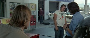 Movie still from “Two-Lane Blacktop” (1971), directed by Monte Hellman – Two men and a woman are standing in a garage; Medium shot, Over the shoulder angle
