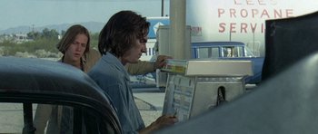 Movie still from “Two-Lane Blacktop” (1971), directed by Monte Hellman – A man sitting in a chair next to a gas pump; Medium shot, Over the shoulder angle