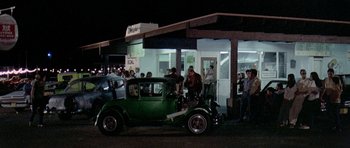 Movie still from “Two-Lane Blacktop” (1971), directed by Monte Hellman – An old car parked in front of a building with people standing around it; Wide shot, High angle