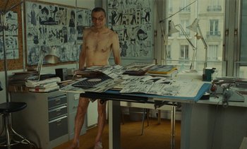 Movie still from “Ulysse” (1983), directed by Agnès Varda – A man standing in front of a table with papers on top of it; Wide shot, Low angle