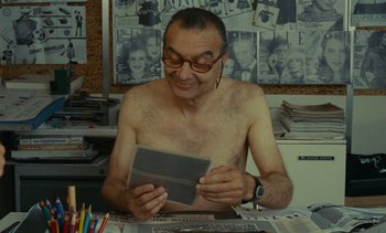 Movie still from “Ulysse” (1983), directed by Agnès Varda – A shirtless man sitting at a table with a book; Medium shot, Over the shoulder angle
