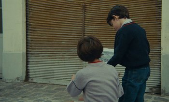Movie still from “Ulysse” (1983), directed by Agnès Varda – Two young boys standing next to each other on the sidewalk; Medium shot, Over the shoulder angle