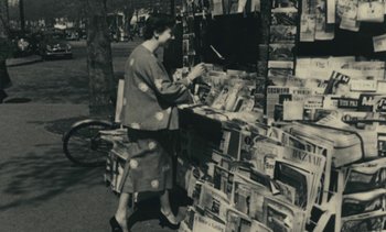 Movie still from “Ulysse” (1983), directed by Agnès Varda – An old black and white photo of a woman standing in front of a pile of newspapers; Medium shot, High angle