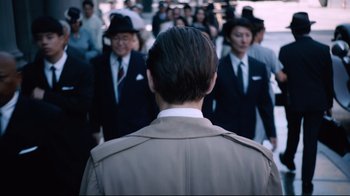 Movie still from “Unbroken: Path to Redemption” (2018), directed by Harold Cronk – A man in a trench coat is standing in front of a group of people; Medium shot, Over the shoulder angle