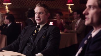 Movie still from “Unbroken: Path to Redemption” (2018), directed by Harold Cronk – A man in a suit and tie sitting in front of a table; Medium shot, Over the shoulder angle