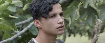 Movie still from “Under the Fig Trees” (2021), directed by Erige Sehiri – A young man with black curly hair looking to his left; Close Up shot, Over the shoulder angle