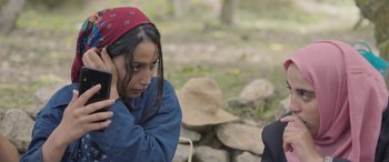 Movie still from “Under the Fig Trees” (2021), directed by Erige Sehiri – A young woman wearing a red hat sitting in front of a stone wall; Close Up shot, Over the shoulder angle