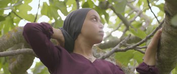 Movie still from “Under the Fig Trees” (2021), directed by Erige Sehiri – A young woman wearing a head scarf standing in a tree; Close Up shot, Low angle