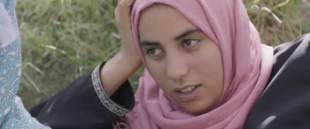 Movie still from “Under the Fig Trees” (2021), directed by Erige Sehiri – A woman in a pink head scarf sitting in the grass; Close Up shot, Over the shoulder angle