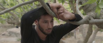 Movie still from “Under the Fig Trees” (2021), directed by Erige Sehiri – A man holding onto a tree branch while wearing a black t - shirt; Close Up shot, Low angle