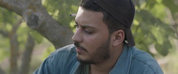Movie still from “Under the Fig Trees” (2021), directed by Erige Sehiri – A person wearing a hat with a pencil in his head; Close Up shot, Low angle