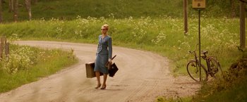 Movie still from “Under the Sun” (1998), directed by Colin Nutley – A woman in a blue dress carrying suitcases and walking down a dirt road; Wide shot, Low angle
