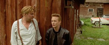 Movie still from “Under the Sun” (1998), directed by Colin Nutley – A young man standing next to another man; Medium shot, Over the shoulder angle