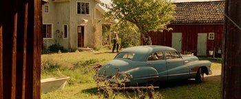 Movie still from “Under the Sun” (1998), directed by Colin Nutley – An old blue car parked in a yard with a person walking by; Extreme Wide shot, Low angle