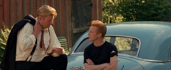 Movie still from “Under the Sun” (1998), directed by Colin Nutley – Two men sitting next to each other in front of an old car; Medium shot, Over the shoulder angle
